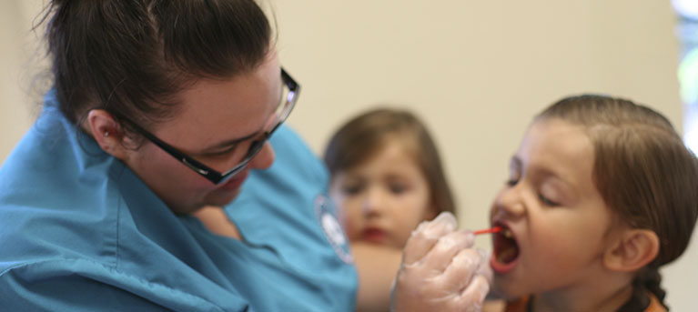 An image of a female dentist in blue scrubs doing an inspection of a young girls mouth with some dental tools